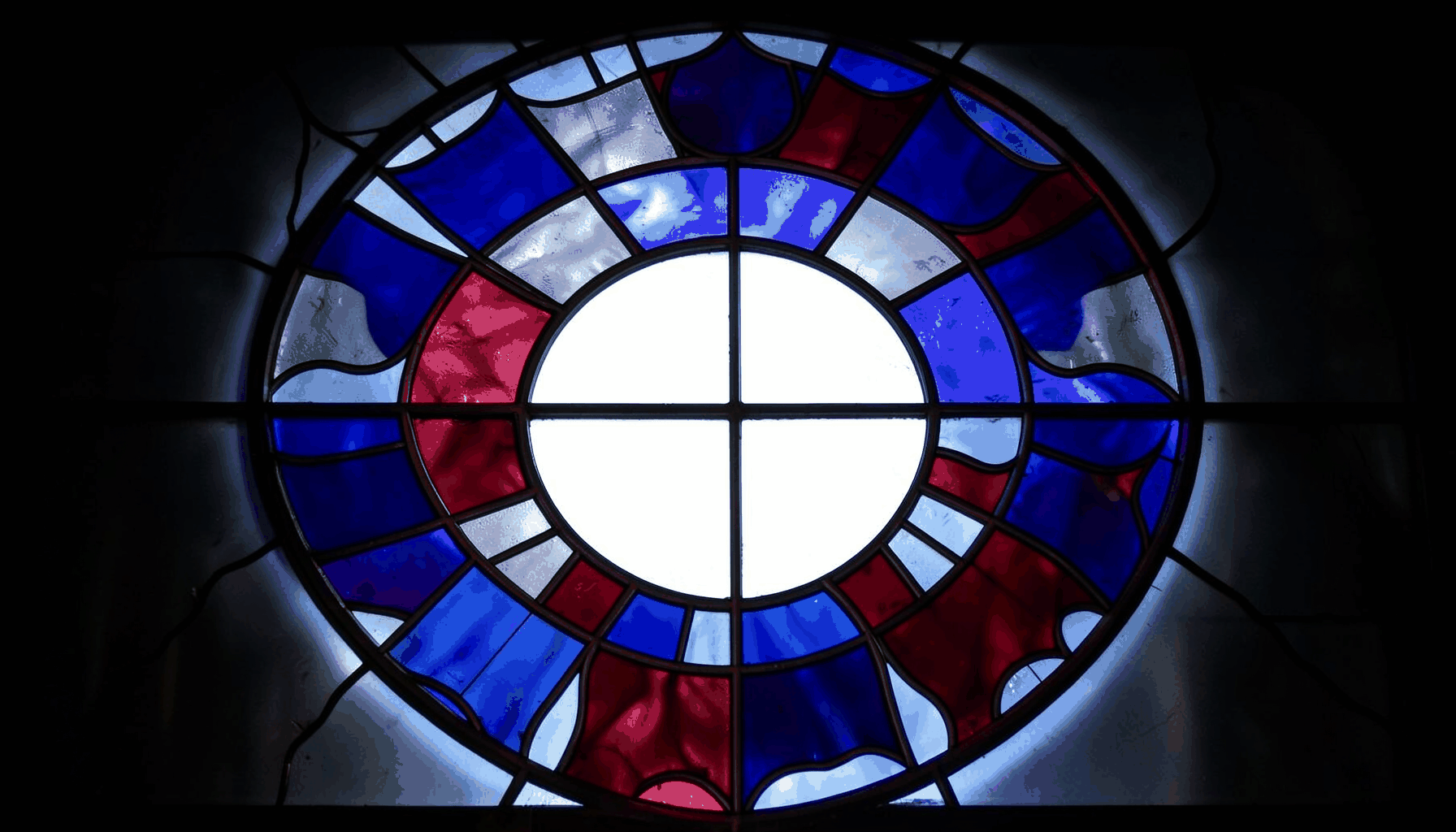 Stained glass composition in bold leaded segments showing a ring of shield shapes in deep blue, steel grey and crimson red, arranged like a globe of banners representing many nations, calm and unified mood